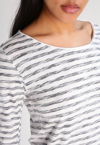 Gestreiftes Langarmshirt in Weiß und Schwarz, mit horizontalen wellenförmigen Linien, einem weiten Ausschnitt und einer weichen Stofftextur.