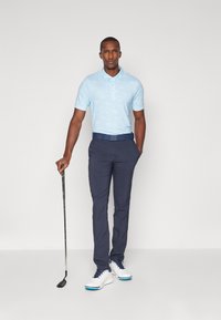 Helles blaues Polohemd mit kurzen Ärmeln und subtilen Mustern, kombiniert mit dunkelblauen Hosen. Weiße Golfschuhe mit blauen Akzenten und einem dunklen Gürtel. Golfclub in der Hand.