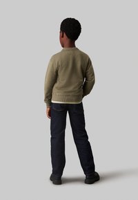 Felpa verde oliva con orlo a coste, abbinata a jeans in denim blu scuro. Il ragazzo è di spalle e indossa scarpe nere.