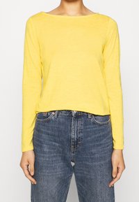 Gelbes Langarmoberteil mit weitem Rundhalsausschnitt, aus leichtem Stoff gefertigt. Kombiniert mit hochgeschnittenen, dunkelblauen Jeans.