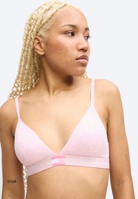 Νεαρή γυναίκα με ανοιχτόχρωμο δέρμα, μακριά ξανθά πλεξούδα μαλλιά, φοράει ένα ανοιχτό ροζ τρίγωνο σουτιέν με τη μάρκα Tommy Hilfiger, κοιτάζει προς τα δεξιά της.