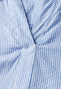 Tessuto a righe blu e bianche con una finitura testurizzata, caratterizzato da una scollatura a V e linee di cucitura angolate. Le righe sono distanziate uniformemente.