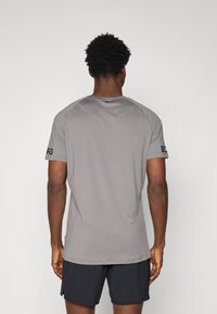 T-shirt de sport gris à manches courtes avec un col rond, présentant des coutures plates et des détails textuels sur la manche gauche. Matériau en mélange de coton, coupe régulière.