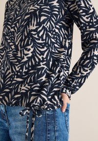 Haut à manches longues bleu marine et blanc avec des motifs de feuilles, en tissu texturé et doté d'un lien au bas, associé à un jean en denim bleu.