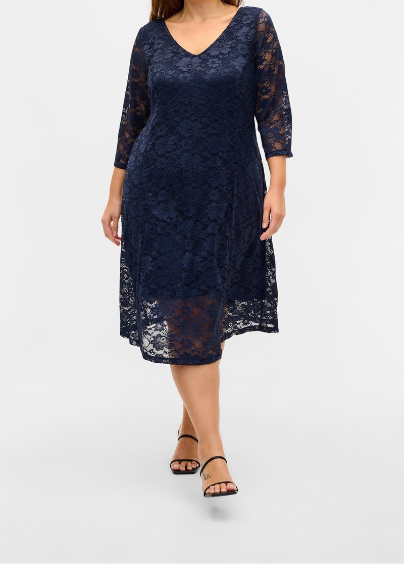Femme portant une robe en dentelle bleu marine longueur genoux avec manches trois-quarts et des sandales noires à lanières sur fond gris.