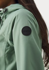 Veste vert clair au toucher doux, avec un logo circulaire noir sur la manche gauche. Comprend une capuche réglable et une fermeture éclair.