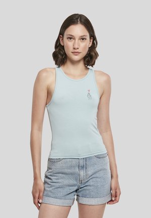Jeune femme aux cheveux courts et bruns porte un haut sans manches bleu clair avec un petit motif main et cœur et un short en jean.