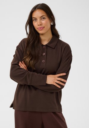 Femme aux longs cheveux bruns portant une chemise marron à manches longues avec col et boutons, les bras croisés, debout devant un fond clair uni.