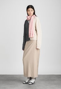 Πλεκτά πουλόβερ με σχέδιο καλωδίων σε σκούρο γκρι και ελεφαντόδοντο, ροζ κασκόλ, κρεμ μαξι φούστα και μαύρες λουστρίνι γόβες με λεπτομέρειες στον αστράγαλο, τοποθετημένα σε ουδέτερο φόντο.