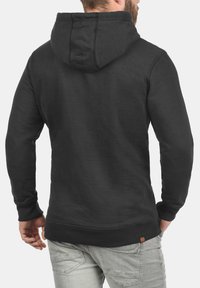 Sudadera con capucha negra de acabado texturizado, puños y dobladillo acanalados. Presenta una costura central en la parte posterior y un pequeño parche del logo cerca de la parte inferior.