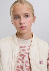 Cremefarbene gesteppte Weste mit einem runden, rosa Logo mit Totenkopf und gekreuzten Knochen, darüber getragen ein hellrosa gemusterter Pullover.