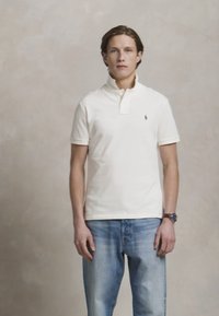 Νεαρός άνδρας που φοράει ένα λευκό polo πουκάμισο και μπλε τζιν, στέκεται μπροστά σε έναν απλό μπεζ φόντο, στραμμένος προς τα εμπρός.