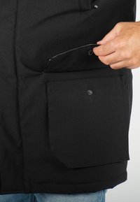 Veste noire avec une poche boutonnée. Le matériau semble lisse, avec des lignes simples et un design épuré. Une main s'approchant de la poche.