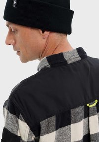 Gorro negro, camisa de franela a cuadros en blanco y negro con una parte superior negra, que presenta un acento amarillo cerca del escote.