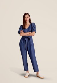 Denim jumpsuit in diepblauw met korte pofmouwen, getailleerde taille en wijde benen. Gestyled met een stoffen strik en witte sneakers.