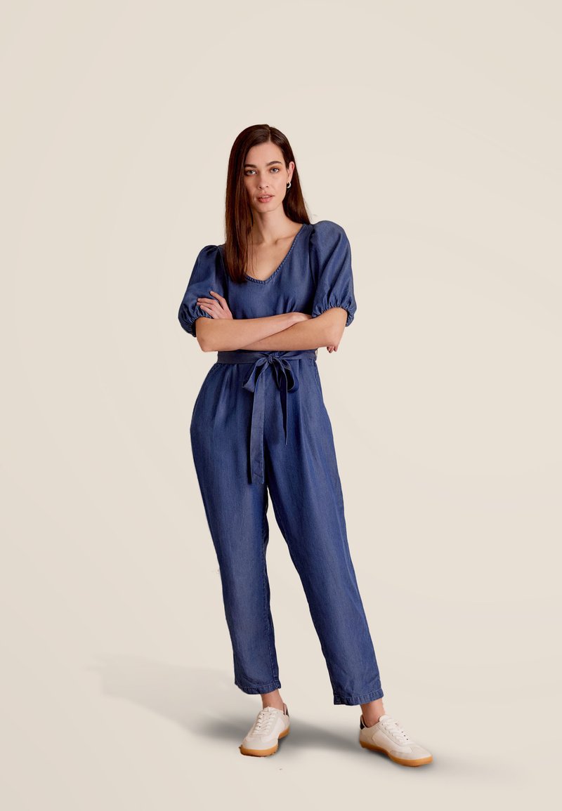 Denim jumpsuit in diepblauw met korte pofmouwen, getailleerde taille en wijde benen. Gestyled met een stoffen strik en witte sneakers.