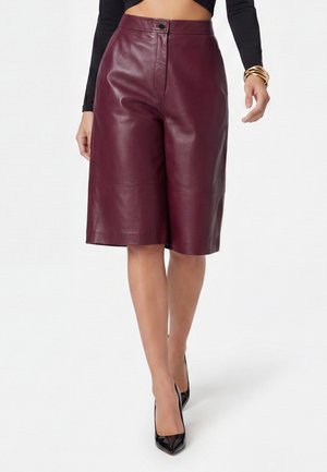 Woman wearing knee-length burgundy leather shorts, black long-sleeve crop top, black pointed high heels, and gold bracelets on one wrist.