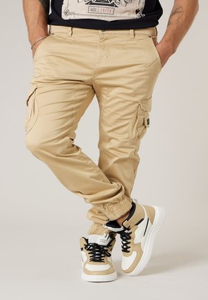 Homme debout, les mains dans les poches, portant un pantalon cargo beige et des baskets beige et blanches avec des lacets noirs sur un fond clair.