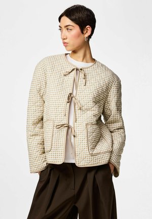 Žena s krátkými vlasy, oblečená v béžovém károvaném prošívaném kabátu se zavazováním na šňůrky a tmavě hnědých kalhotách, s rukama v kapsách, hledící doleva.