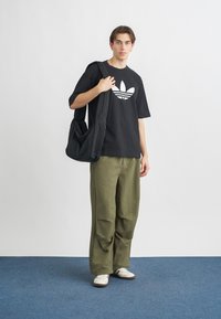Black cotton t-shirt with white logo, olive green baggy pants, and white shoes with black stripes. Model holding a black shoulder bag.