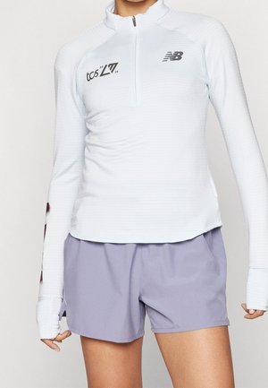 Person iført en hvid atletisk trøje med kvart lynlås og logoer samt lyse lilla shorts, stående med den ene hånd delvist synlig.