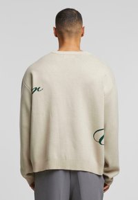 Beige oversized trui met groene geborduurde accenten, geribbelde manchetten en zoom, en een ronde hals. Zachte stoftextuur met een ontspannen pasvorm.
