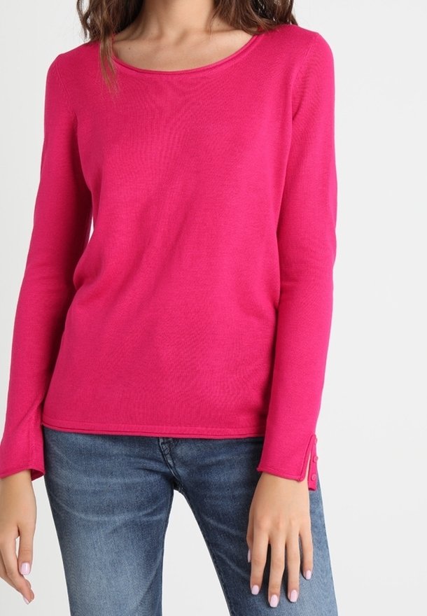 Femme portant une chemise rose vif à manches longues avec des détails de boutons aux poignets et un jean bleu sur fond blanc.