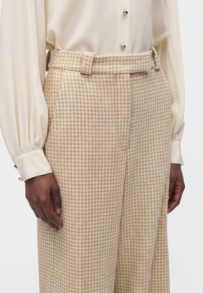Personne portant un pantalon taille haute beige et blanc à motif pied-de-poule et un chemisier crème à manches longues avec des boutons argentés.