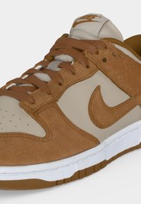 Κοντινό πλάνο ενός αθλητικού παπουτσιού Nike με καφέ σουέτ επενδύσεις, μπεζ δερμάτινη βάση, καφέ κορδόνια και λευκή και καφέ σόλα.