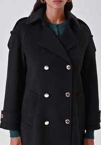 Cappotto nero doppiopetto realizzato in tessuto strutturato, con bottoni dorati, tasche laterali e colletto a nota. I dettagli includono particolari sui polsini.