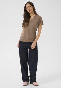 T-shirt marron à manches courtes et col en V associé à un jean bleu foncé à jambes larges et à des baskets rose clair, sur un fond uni clair.