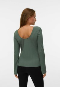 Langemmentongen top in gedempt groen, met een ronde rug en een gestructureerde stof. Gecombineerd met zwarte onderkleding, wat een aangesloten silhouet oplevert.