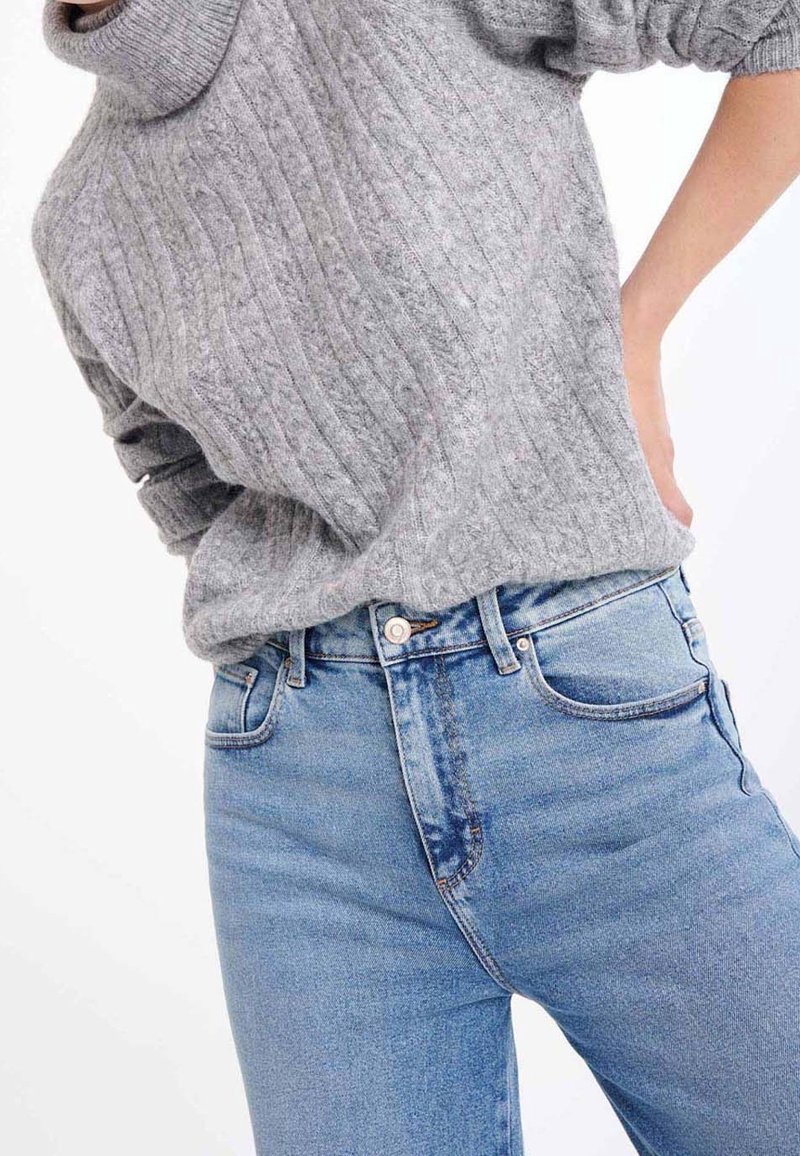 Szary prążkowany sweter z wysokim kołnierzem w zestawieniu z jasnoniebieskimi, wysokim stanem jeansami o minimalistycznych przeszyciach i klasycznym fasonie z pięcioma kieszeniami.