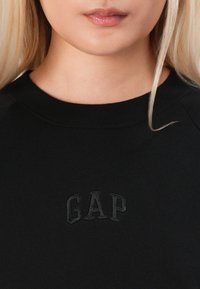 Close-up van een persoon die een zwart sweatshirt draagt met een klein zwart reliëf GAP-logo op de borst en zichtbare lippen en hals.