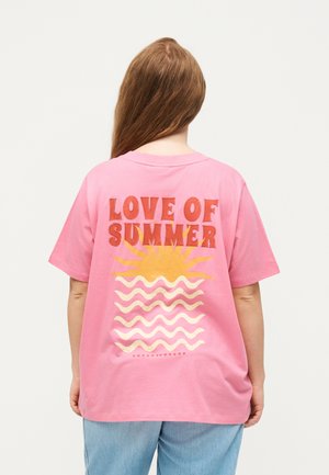 Person med langt brunt hår iført en lyserød T-shirt med teksten "LOVE OF SUMMER", sol- og bølgedesign på ryggen, kombineret med lyseblå jeans.