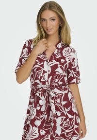 Abito camicia a fiori rosso e bianco con maniche corte, chiusura frontale a bottoni e vita legata, realizzato in tessuto leggero.