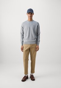 Pull gris clair, chinos beiges, mocassins marron et casquette bleu marine. Le pull présente des poignets côtelés et un détail de logo sur la poitrine. Texture lisse dans l'ensemble.
