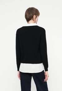 Une personne avec les cheveux bruns courts porte un pull noir sur une chemise blanche et un pantalon sombre, de dos contre un fond blanc uni.