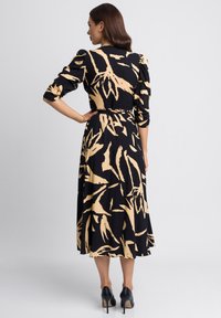 Czarna sukienka w beżowy kwiatowy wzór, z rękawami w 3/4, zwężoną talią i zwiewną spódnicą, w połączeniu z czarnymi szpilkami.