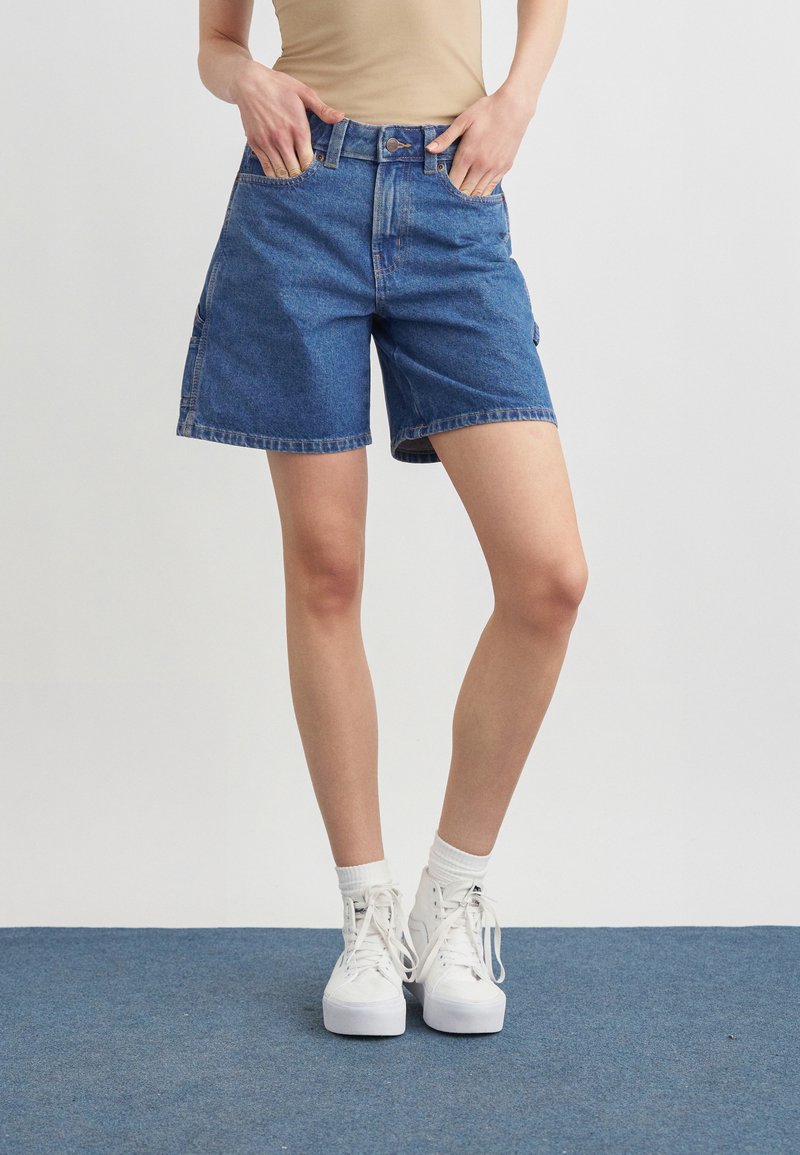 Femme portant un short en jean bleu mi-long, un haut beige, des chaussettes blanches et des baskets à plateforme blanches sur un fond uni.
