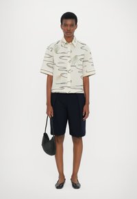 Kortermet skjorte med en lys beige bakgrunn og abstrakte grønne og brune mønstre, parret med marineblå shorts og en svart teksturert håndveske.