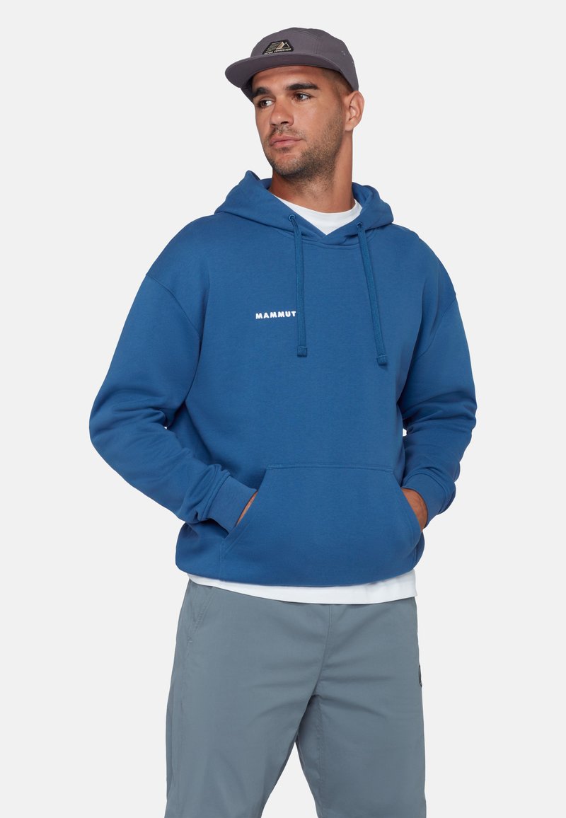 Man die een blauwe Mammut-hoodie, een grijze pet en lichtgrijze broek draagt, met zijn handen in de zak van zijn hoodie, kijkend naar de zijkant.