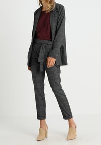 Veste à carreaux gris avec un pantalon assorti à ceinturer, associée à un haut bordeaux. Des chaussures à talons en nubuck beige complètent la tenue.