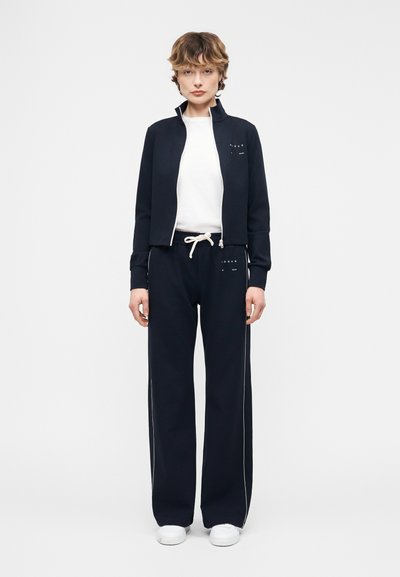 Personne debout, faisant face à l'avant, portant une veste noire zippée, une chemise blanche, un pantalon noir à jambes larges avec cordon de serrage, et des baskets blanches sur fond blanc.