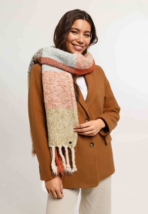 Smiling woman wearing a brown coat, multicolored thick scarf, and light-colored pants, standing against a plain background.