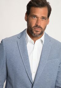 Blazer azzurro chiaro con texture, colletto a lancia, chiusura a un bottone e dettaglio del fazzoletto da taschino, indossato sopra una camicia bianca con colletto.