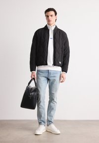 Chaqueta bomber acolchada negra con cremallera, capa de forro polar blanca debajo, jeans desgastados azul claro, zapatillas blancas, llevando una bolsa de deporte negra.