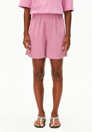 Personne portant un short à taille élastique violet clair et un haut assorti, debout pieds nus dans des sandales noires et blanches sur fond blanc.