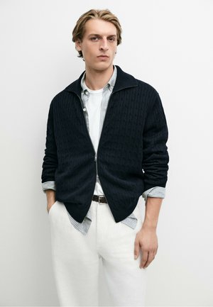 Jeune homme portant un pull zippé noir sur une chemise rayée et un pantalon blanc, debout les mains dans les poches devant un fond uni.