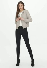 Jaqueta cinza clara com fecho éclair e capuz, usada sobre uma t-shirt branca, combinada com jeans skinny pretos e botas de tornozelo. Texturas suaves, design casual.